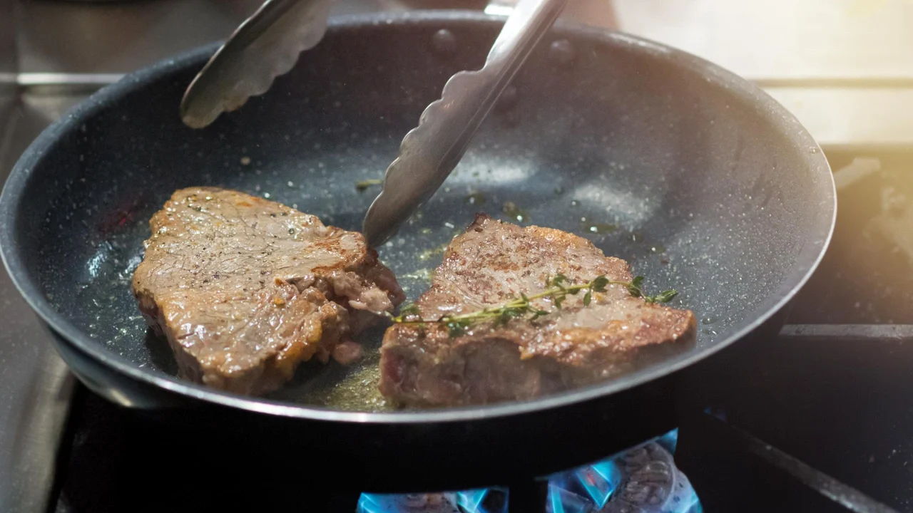Due bistecche in cottura in padella con rametto di timo e pinza da cucina sul fuoco a gas